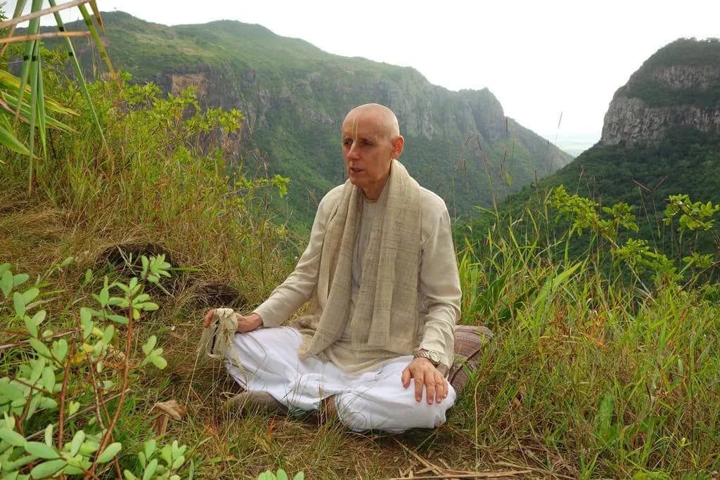 A devotee chanting japa, seated on a green hillside